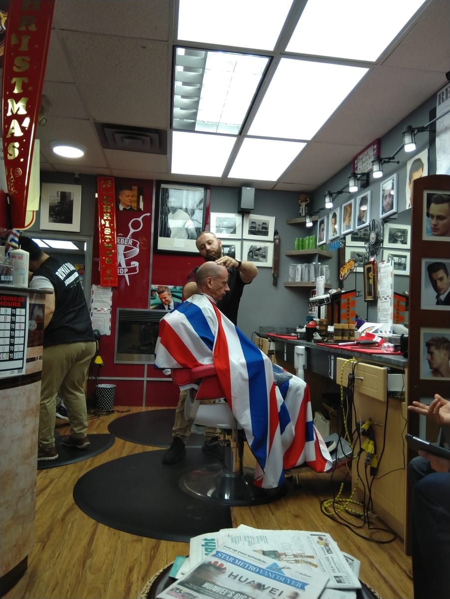 A barber cutting a client wrapped in a Union Jack cape, framed portraits along the wall